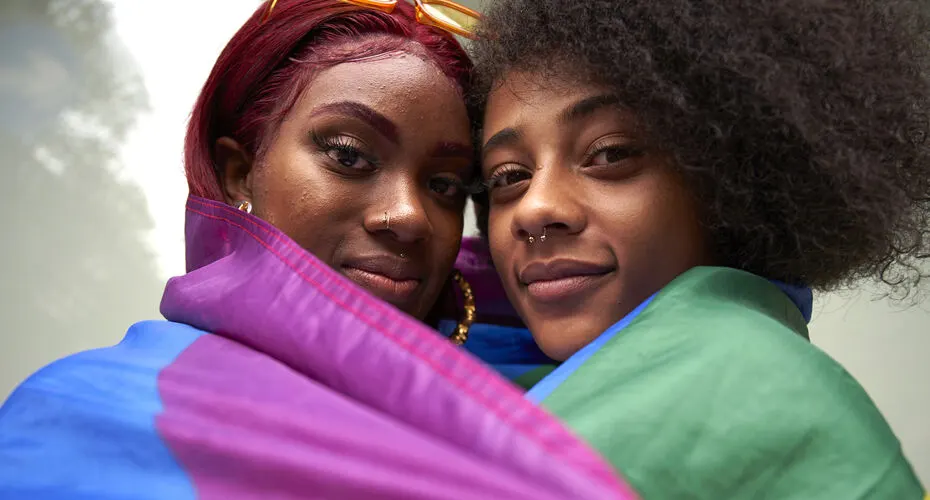 Two people wrapped in an LGBTQ+ flag looking at the camera.